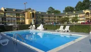 The image shows a clear outdoor swimming pool with lounge chairs in front of a multi-story motel complex on a sunny day.
