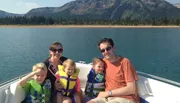 A family of two adults and three children is enjoying a sunny day on a boat with a scenic mountainous backdrop.