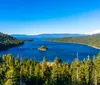 The image shows a panoramic view of a serene blue lake surrounded by dense green coniferous forests with a small island in the center under a clear blue sky