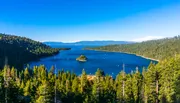 The image shows a panoramic view of a serene blue lake surrounded by dense green coniferous forests with a small island in the center, under a clear blue sky.