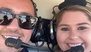 Two smiling people wearing aviation headsets are taking a close-up selfie, with reflective sunglasses revealing someone's hand holding the camera.