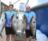 Two smiling men are proudly displaying large fish they appear to have caught standing on a dockside with a boat in the background